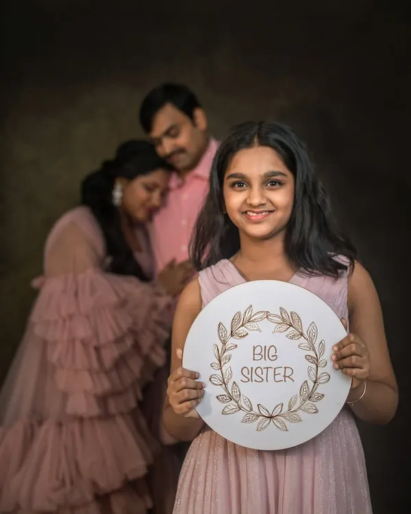 A young girl proudly holds a "Big Sister" sign, with her expecting parents softly focused in the background. This is a creative and heartwarming way to announce a new addition to the family.