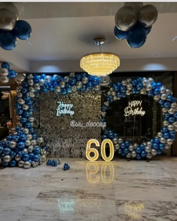 An expansive and luxurious setup for a 60th birthday, featuring two shimmer walls, double neon signs, and a large balloon arch in blue and silver, perfect for a grand celebration.