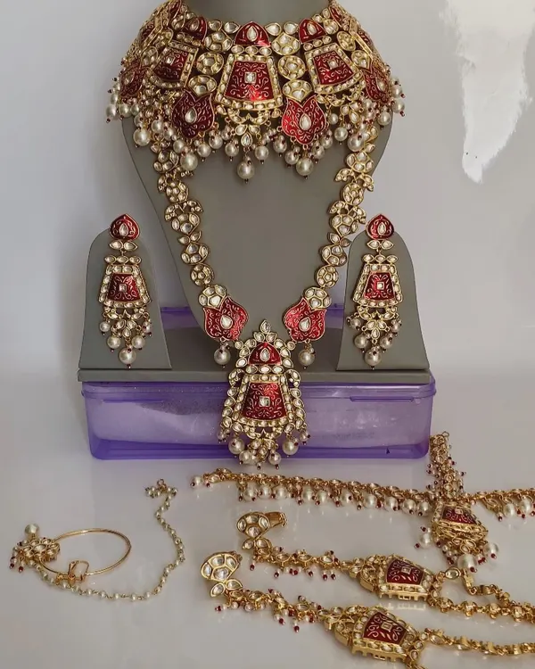 A complete bridal ensemble featuring a red meenakari choker and a longer kundan necklace. This set also includes a mathapatti, hathphool, and nath, all designed to create a cohesive and stunning bridal look.