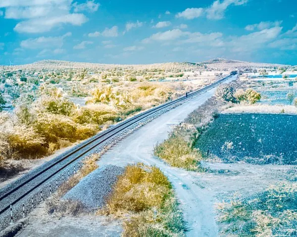 An infrared photograph of a railway line cutting through the landscape. The foliage turns a golden yellow and the sky a vibrant blue, creating a surreal, false-color world.