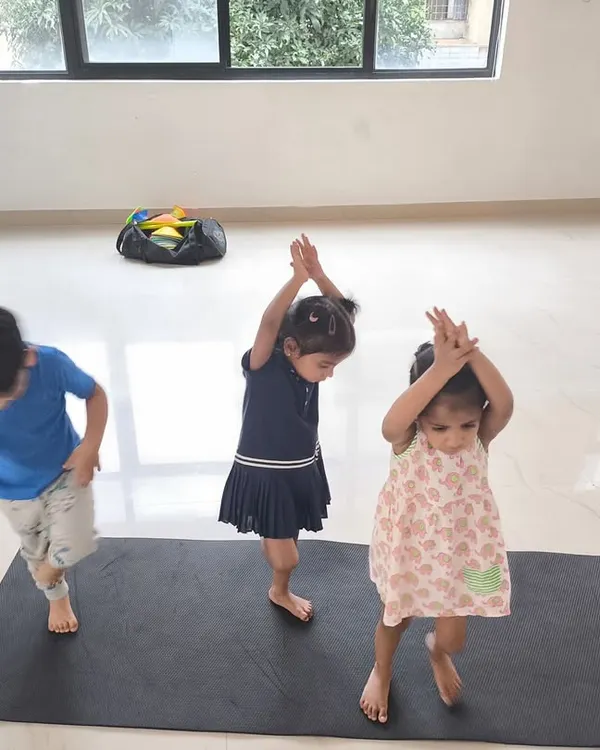 Three young children attempt a gymnastics pose during our indoor fitness class. We use playful imitation to teach basic movements, making learning fun and engaging for toddlers.