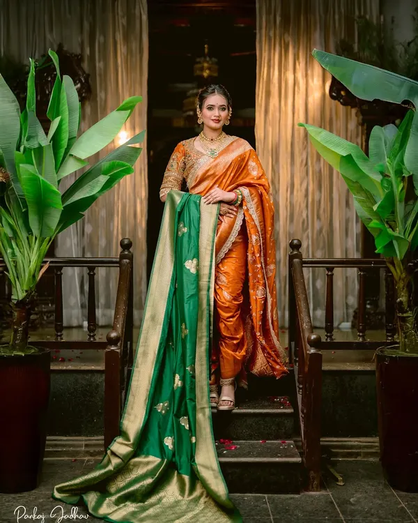 A portrait of pure elegance. The bride stands on a staircase, her vibrant orange saree and contrasting green shawl creating a stunning visual against the traditional decor.