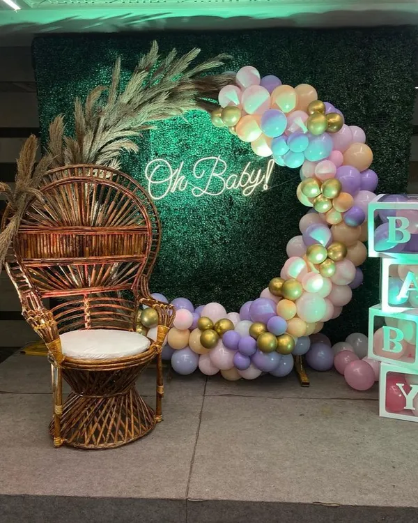 A popular baby shower decoration featuring a bohemian-style wicker chair for the mom-to-be. The setup includes a circular balloon arch in pastel colors against a green hedge wall, an 'Oh Baby!' neon sign, and 'BABY' blocks.