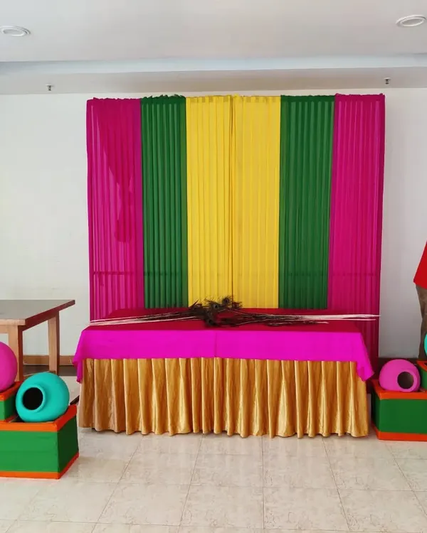 My team setting up a colorful pooja corner for a Janamashtmi celebration. The backdrop uses bright pink, green, and yellow drapes, with decorative matkis placed on either side of the main table.