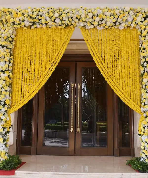 A stunning entrance for a daytime ceremony, featuring pillars covered in white and yellow flowers and curtains made of traditional marigold garlands.
