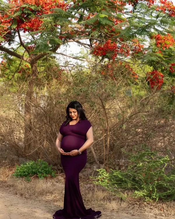 Motherhood unplugged, surrounded by nature's beauty. The vibrant red flowers of the Gulmohar tree perfectly complement the deep purple of her gown in this serene outdoor portrait.