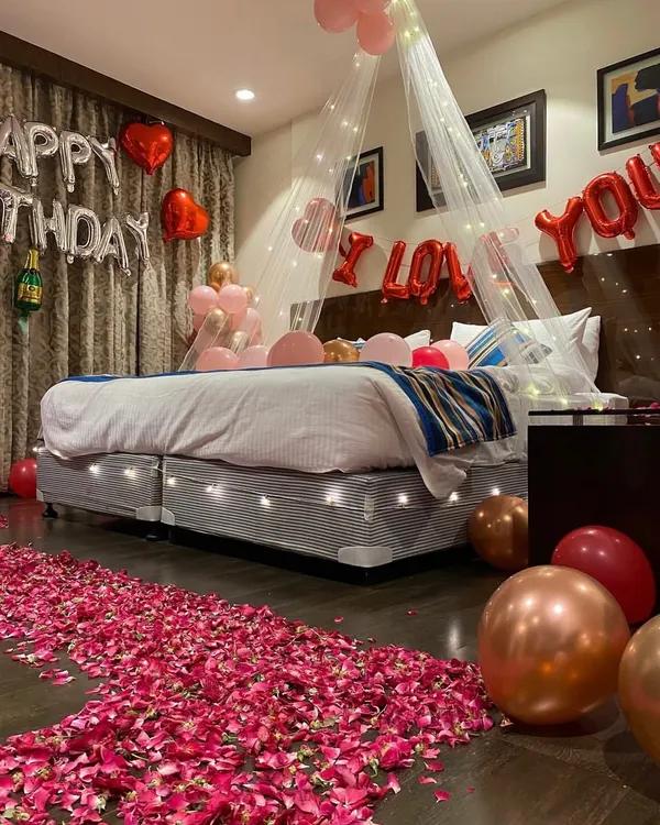 A complete romantic birthday surprise decoration. This room setup includes a bed canopy with fairy lights, a rose petal pathway, 'Happy Birthday' and 'I Love You' foil balloons, and a mix of regular and heart-shaped balloons.