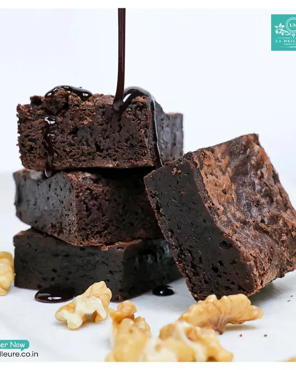 A stack of our fudgy walnut brownies with a decadent stream of chocolate sauce being poured over them. You can see the rich, dense texture in every square.