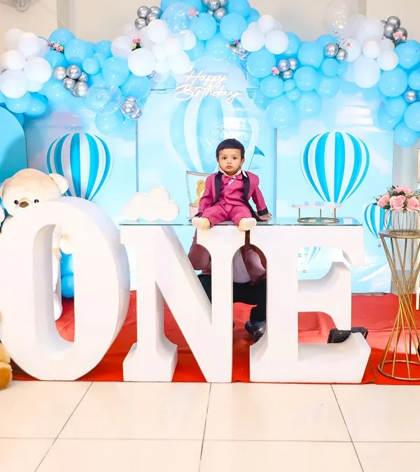 A grand first birthday celebration with a hot air balloon theme. The setup features large 'ONE' letters, a beautiful blue and white balloon arch, and adorable teddy bear accents, creating a picture-perfect scene for the birthday boy.