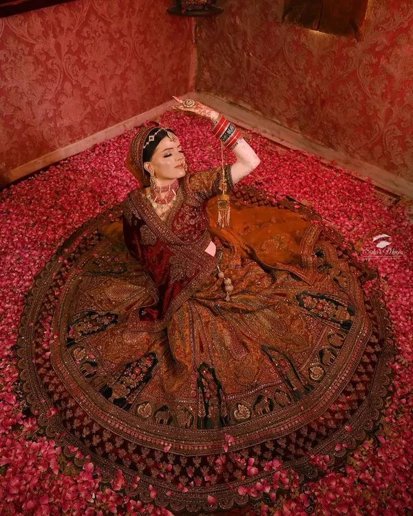 This is the ultimate royal bride moment. I designed this deep red bridal lehenga with an expansive, full-circle skirt covered in intricate, traditional embroidery. The way the skirt spreads out among the flower petals shows the grand silhouette I can create for your wedding day look.