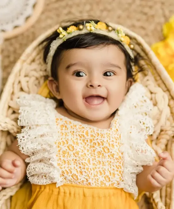 A happy 6-month-old baby girl with a bright, infectious smile. This close-up from her sitter session captures her personality perfectly in a cheerful yellow-themed setup.