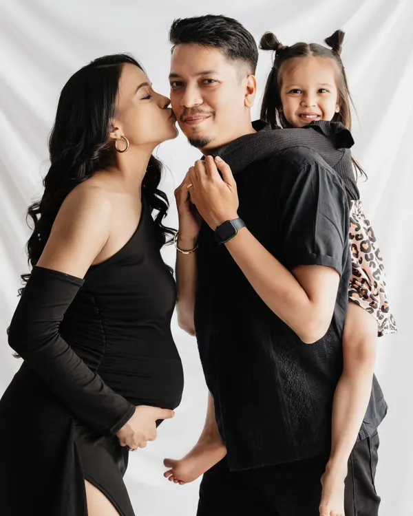 A beautiful family portrait celebrating their journey to becoming a family of four. The mother's baby bump is the focus, while the father and daughter share a fun, loving moment, all captured in a classic studio setting.