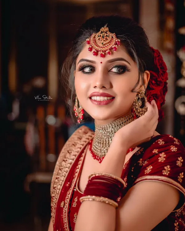 A stunning close-up of a bride in her red wedding lehenga. Her elegant pose, beautiful smile, and the intricate details of her maang tikka and choker necklace are the focus of this classic bridal portrait.
