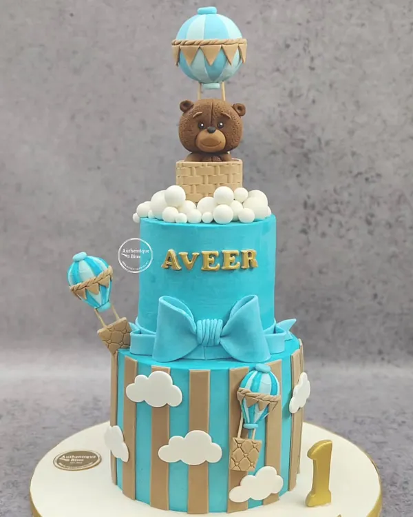 A grand two-tiered hot air balloon cake for a first birthday. This impressive non-fondant cake features a cute teddy bear in a basket, surrounded by clouds and smaller balloons.