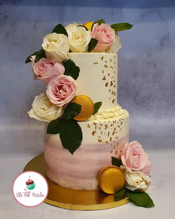 A romantic two-tier cake featuring a soft pink ombre effect on the bottom layer and a cascade of fresh pink and white roses. Gold macarons and delicate gold leaf add a touch of luxury to this wedding cake design.