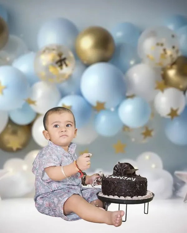A classic cake smash setup for a first birthday. The baby is ready to dig into the cake against a backdrop of blue and gold balloons.
