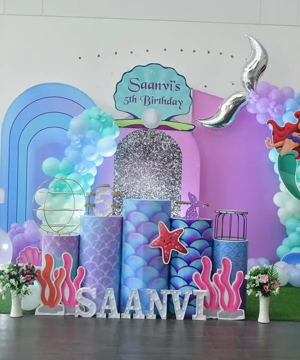 This is a complete mermaid theme I created for Saanvi's 5th birthday. The setup includes a shimmering backdrop, custom-printed pedestals with a scale pattern, balloon arches in ocean colors, and light-up marquee letters to create an immersive underwater world.