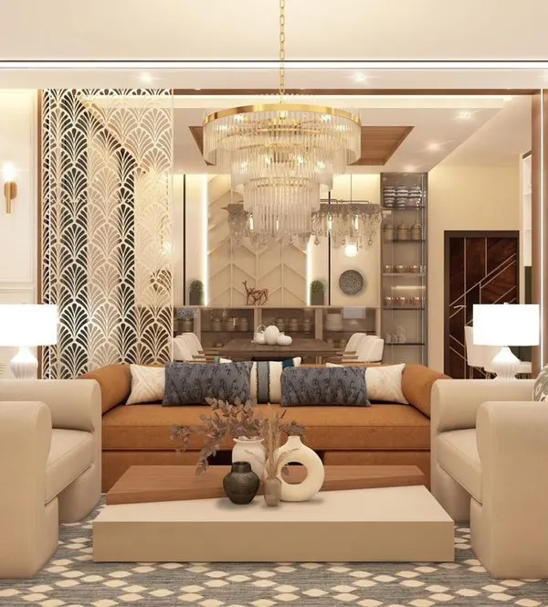 This luxurious living room features a double-height ceiling and a grand chandelier, creating an immediate sense of scale and opulence. The neutral color palette and elegant furnishings are complemented by a geometric jaali screen, adding a touch of traditional Indian craftsmanship to the modern design.
