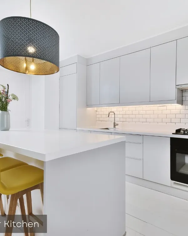 A sleek, all-white modular kitchen with an island that doubles as a breakfast bar. The design features handle-less cabinets, subway tile backsplash, and under-cabinet lighting for a bright and modern workspace.