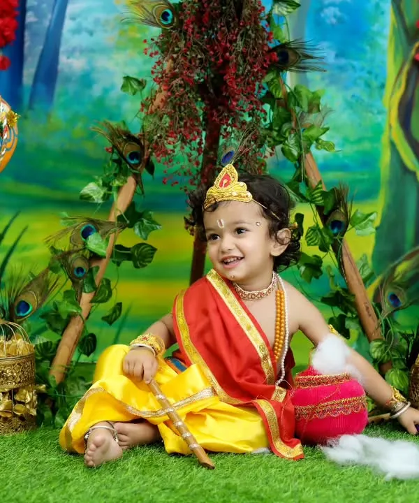 A beautiful portrait of a toddler dressed as Lord Krishna, set against a lush, painted forest backdrop. The detailed costume and golden props create a truly divine image.
