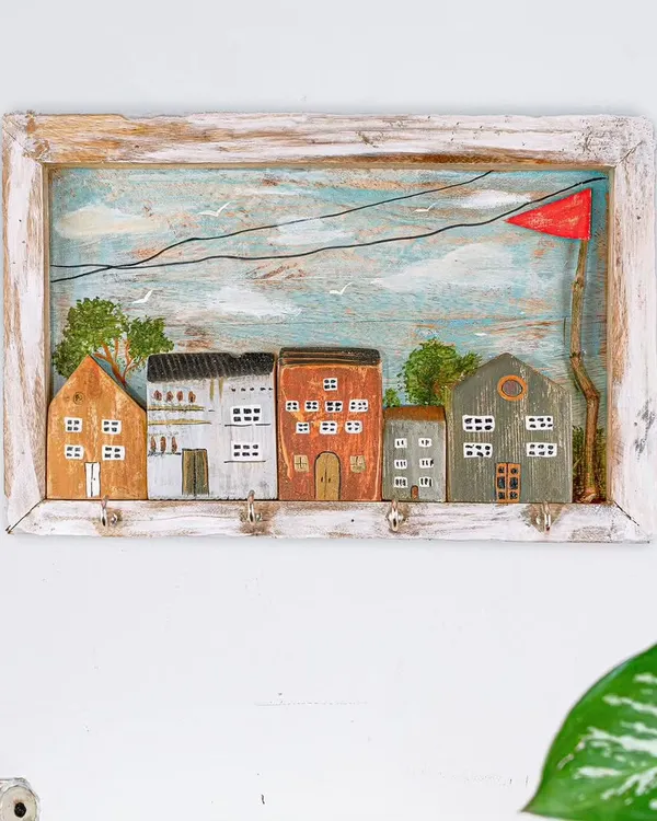 A functional piece of art, this keyholder features a hand-painted scene of miniature houses. It's crafted from wood with a rustic frame, perfect for keeping your keys organized in style.