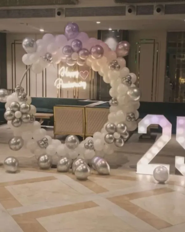 An elegant 25th-anniversary celebration with a white and silver circular balloon arch, a 'Happy Anniversary' neon sign, and large marquee numbers.