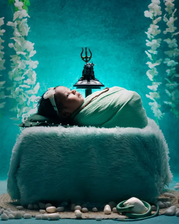 A divine portrait celebrating new life, this Maha Shivratri theme features a peacefully sleeping newborn swaddled in soft green fabric. The setup includes a miniature Trishul and Shivling against a serene turquoise backdrop, creating a truly unique and spiritual memory.