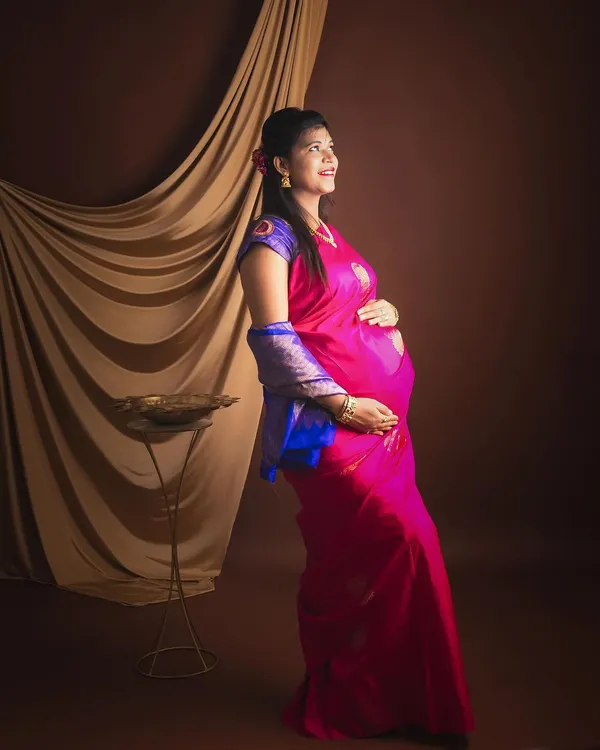 A beautiful maternity portrait of a mother-to-be in a vibrant pink and blue saree, looking down at her bump with a gentle smile.