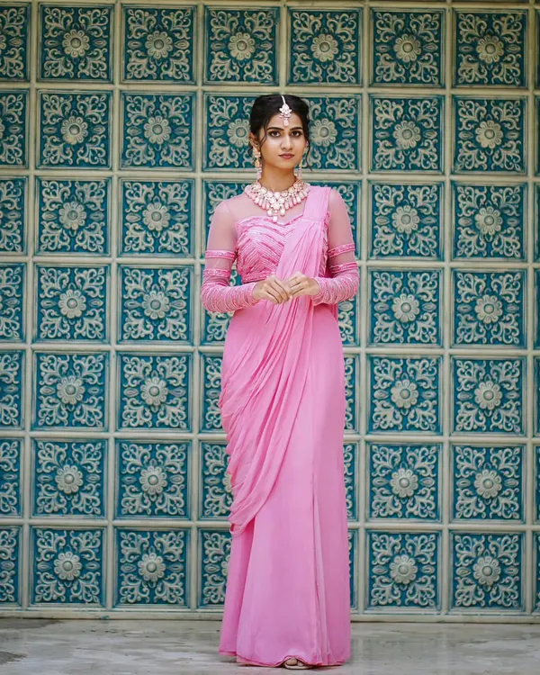 A full-length view of our elegant pink pre-draped saree. The outfit features a stylish blouse with sheer, embellished sleeves and a gracefully draped pallu for a modern silhouette.