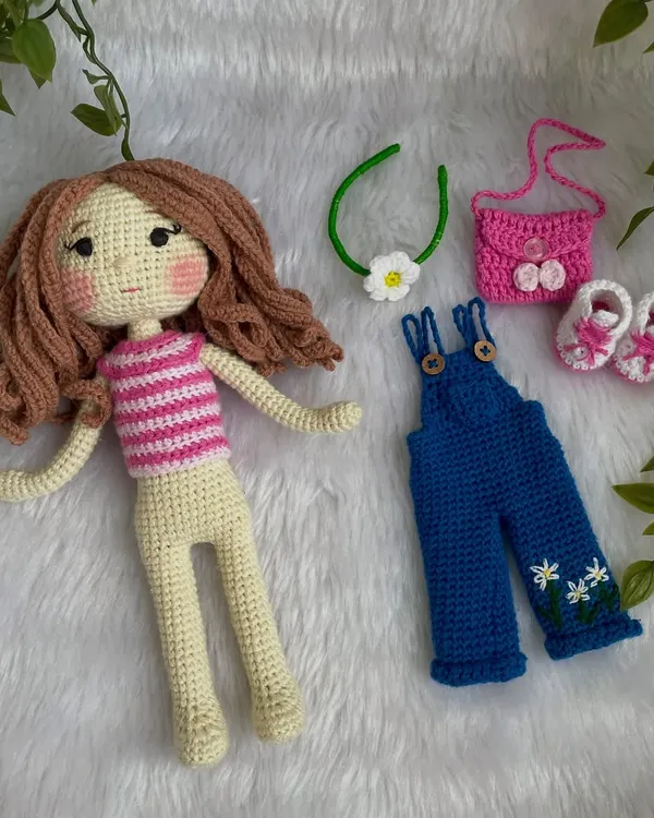 This doll is all ready for playtime with her full set of removable clothes. The set includes a striped top, blue overalls with daisy embroidery, pink shoes, a matching bag, and a flower headband.