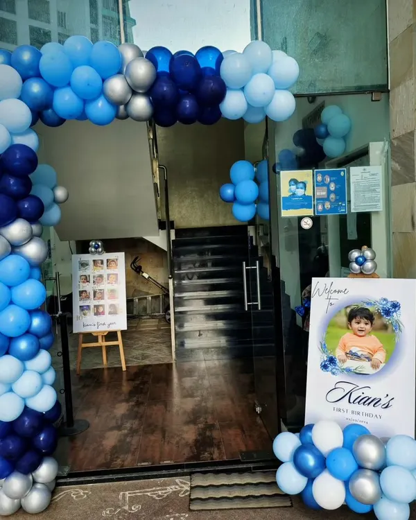 A grand entrance for Kian's first birthday, featuring a full balloon arch in shades of blue and silver. A personalized welcome sign and a milestone photo board greet the guests.