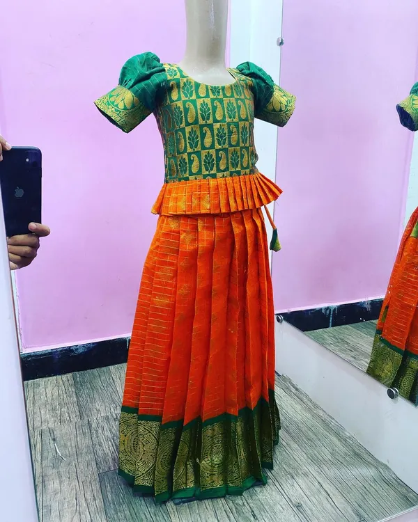 A traditional Paavada blouse set for kids, made with a vibrant orange silk skirt and a green brocade peplum top. The puff sleeves and zari border are classic details for festive wear.