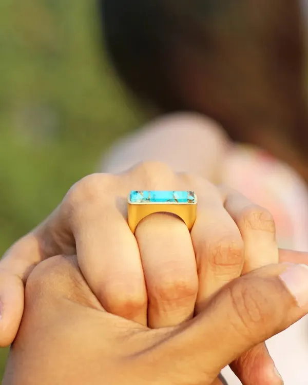 A lifestyle shot that tells a story. By showing the model's hand held by another, the focus on the turquoise statement ring becomes about connection, making it more than just a product photo.