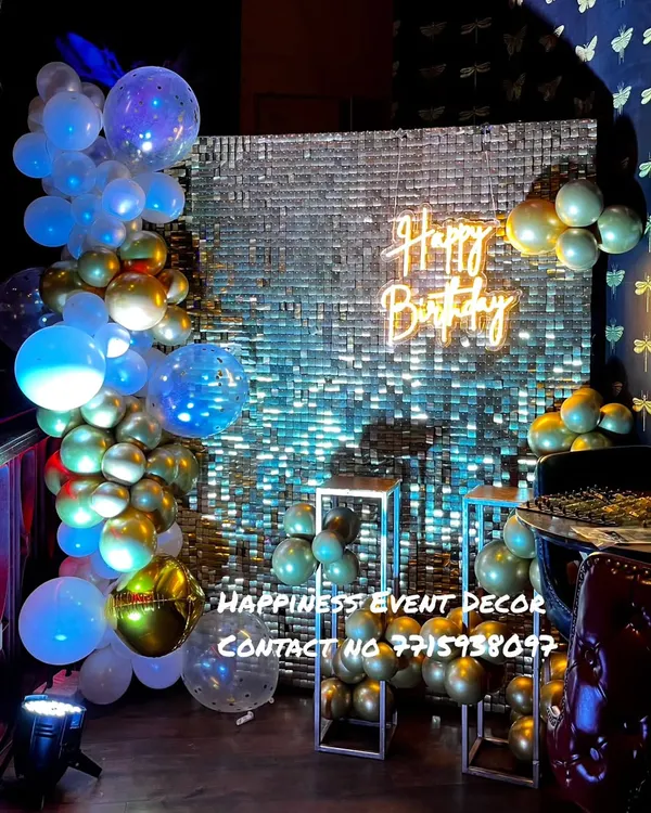 A glamorous birthday setup with a silver sequin backdrop. The decor is enhanced with a balloon arrangement in blue and gold, a 'Happy Birthday' neon sign, and clear plinths for cakes or props.