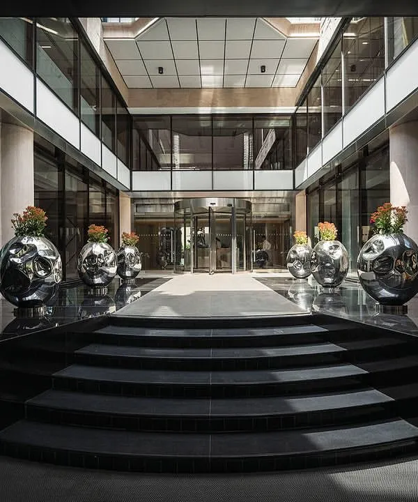 The entrance to 'The Layered Office' features a new canopy and black granite flooring leading to a revolving door. The open-air courtyard is lined with custom stainless steel planters, creating a grand and modern welcome.