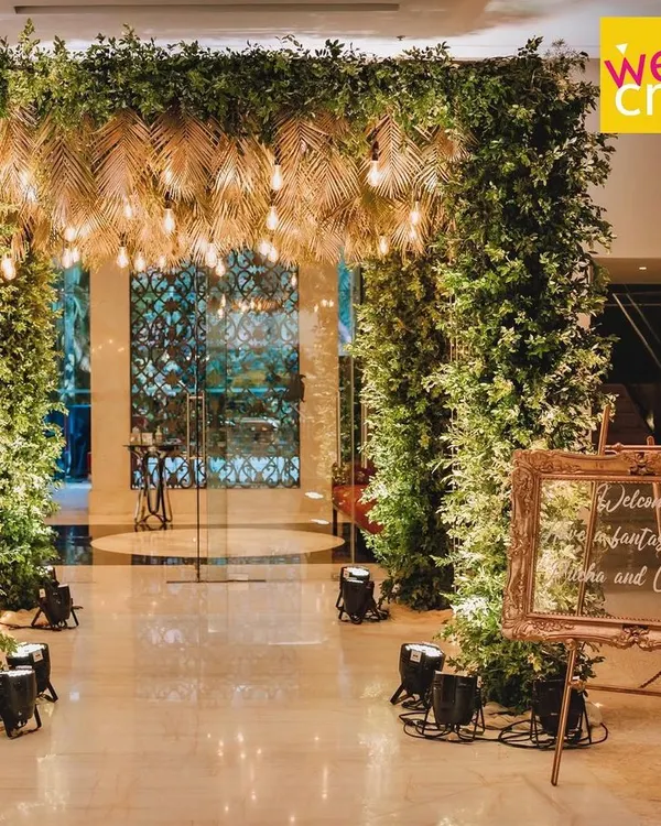 The grand entrance for Rucha and Chinmay's Sangeet night. A lush tropical green archway with a ceiling of golden palm leaves and warm Edison bulbs set the perfect mood.