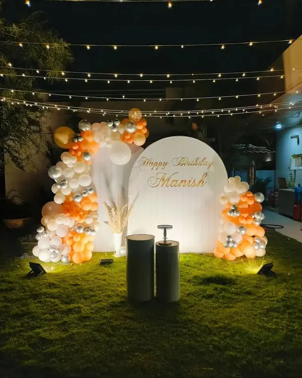 An elegant garden party setup for a birthday. I used a simple white arched backdrop, a balloon arrangement in orange, white, and silver, and overhead string lights to create a sophisticated evening look.