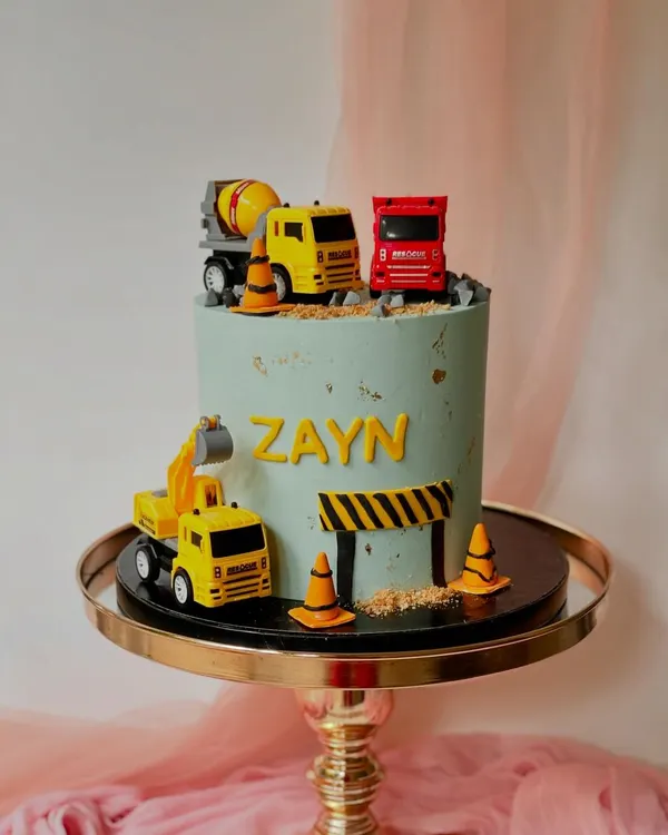 A construction-themed cake with 3D elements. This design features toy trucks and excavators on a pile of "dirt" (crushed cookies) and a road, perfect for a young vehicle enthusiast.