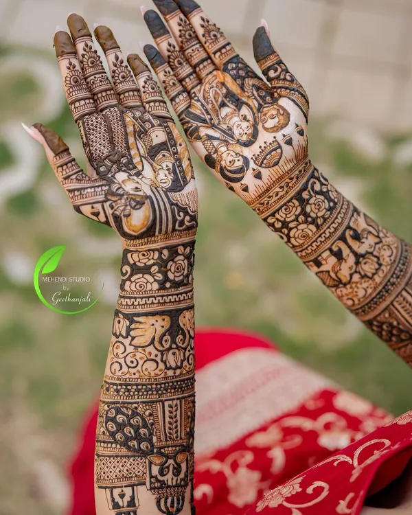 A full view of a stunning bridal mehendi, showcasing intricate patterns from the elbow down. The design features portraits, peacocks, elephants, and beautiful floral bands.