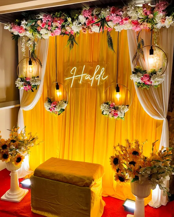 A beautiful and classic Haldi setup. This design features a bright yellow fabric backdrop, a neon "Haldi" sign, and elegant floral hoops with hanging lights.