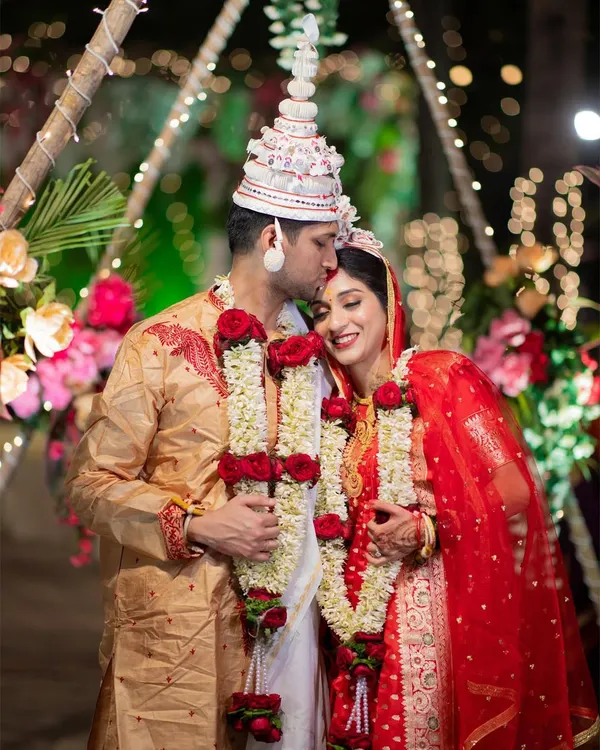 A tender moment from a Bengali wedding, with the groom kissing the bride's forehead. The traditional 'topor' and 'mukut' headwear are beautifully featured.