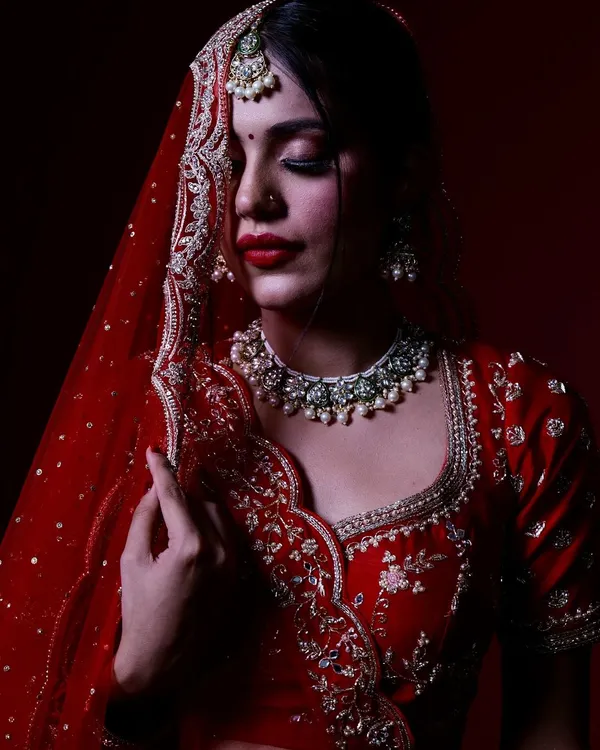 This portrait uses a deep red background and focused lighting to highlight the intricate details of the bridal lehenga and jewelry, creating a classic, elegant look.