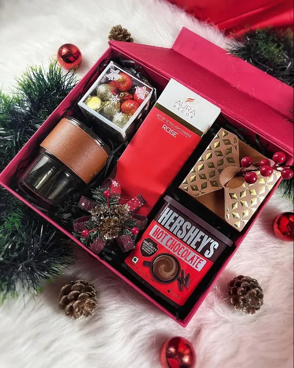 Our 'Holiday Cheer' gift box is filled with everything you need for a cozy Christmas. It includes Hershey's hot chocolate, a rose-scented candle, a glass tumbler, and other festive treats in a vibrant red box.