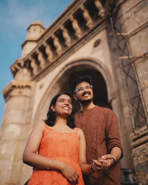 A low-angle shot of Nikit and Anusha looking up towards the sky, with the magnificent Gateway of India arching above them, capturing a moment of shared dreams in their favorite city.