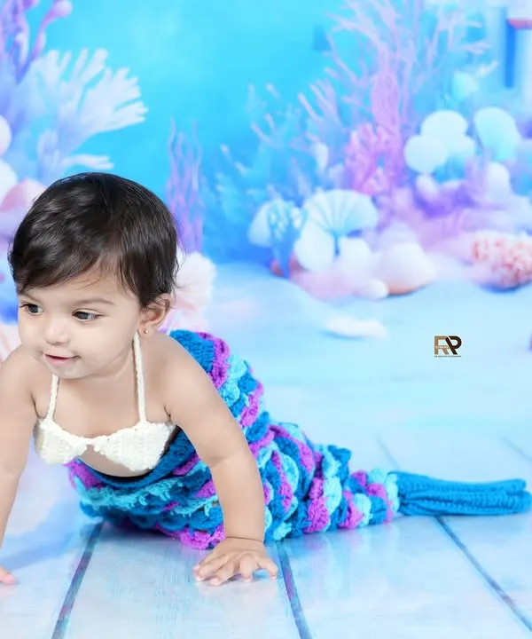 A baby girl dressed as a mermaid crawls across a dreamy, underwater-themed backdrop. The hand-knitted tail and seashell top complete this magical fantasy photoshoot.