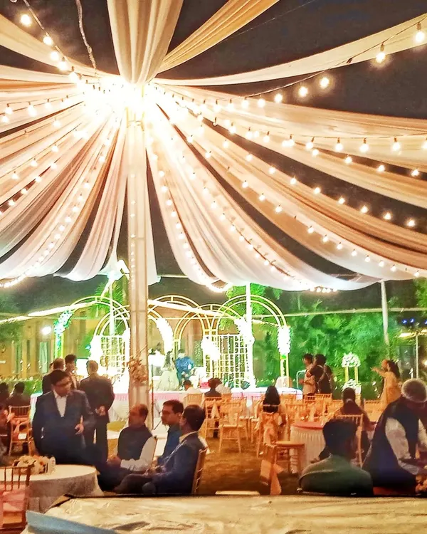 An elegant evening reception setup under a large canopy. The ceiling is draped with cream-colored fabric and lit with strings of Edison bulbs, creating a warm and inviting ambiance.