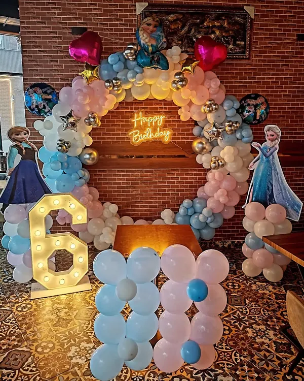 A complete 'Frozen' themed birthday party setup for a 6th birthday. This includes a circular balloon arch in pastel pink, blue, and white, a 'Happy Birthday' neon sign, character cutouts of Elsa and Anna, and a large light-up number '6'.