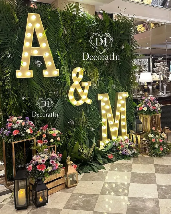 A stunning engagement photo wall featuring a lush backdrop of tropical greenery. The couple's initials, "A & M," in large marquee lights, create a personalized and glowing focal point for pictures.