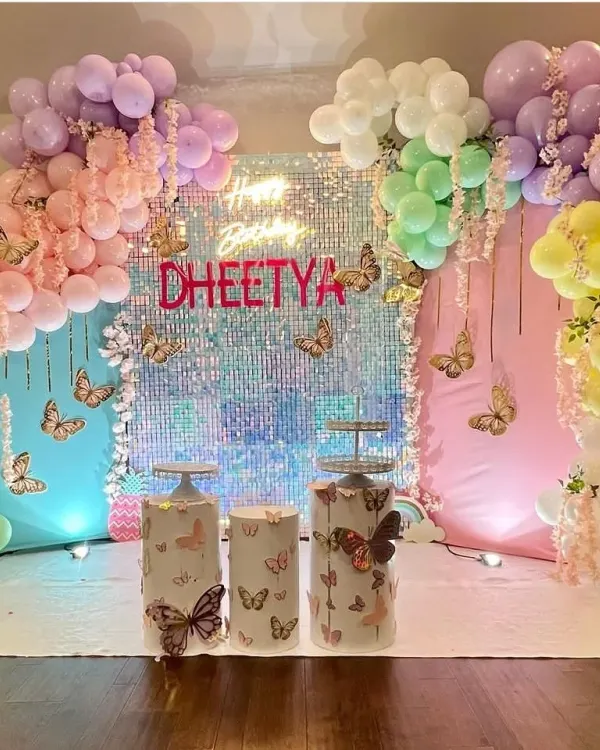 A dreamy butterfly theme with a shimmering sequin backdrop and a rainbow of pastel balloons. This setup for Dheetya's birthday includes custom butterfly-themed pedestals and hanging floral elements for an ethereal feel.