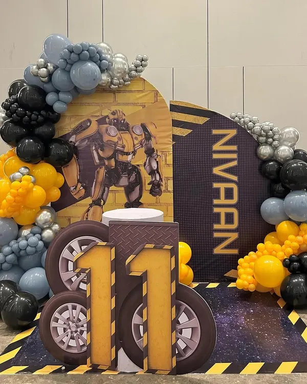 An action-packed Transformers theme for an 11th birthday, featuring a Bumblebee backdrop and a large, custom-made number '11' with a tire-track design. The scene is framed by a balloon arch in signature yellow, black, and silver, ready for an Autobot celebration.